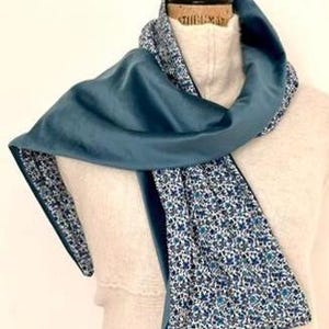 May include: A teal scarf with a floral lining. The scarf is draped over a mannequin, highlighting its smooth texture and contrasting patterns. The floral pattern features small white and blue flowers.