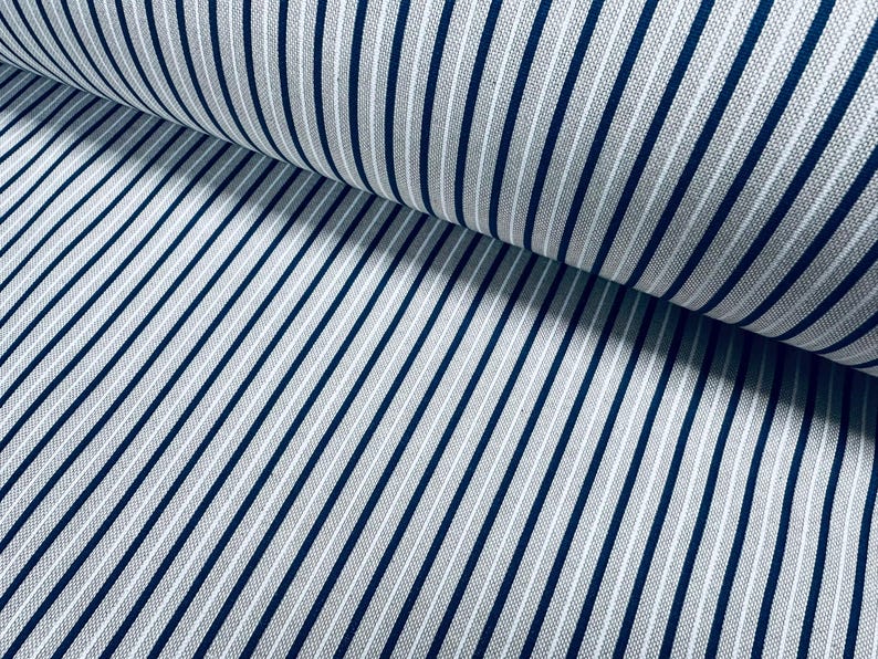 May include: A bolt of fabric featuring a classic pinstripe pattern with thin, dark navy blue vertical lines set against a light gray, textured background. The material is folded over itself, creating a soft curve that highlights the consistent spacing of the stripes across the surface of the textile.