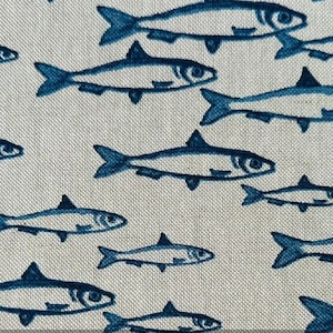 Puede incluir: Primer plano de una tela con un patrón repetitivo de ilustraciones de peces azules sobre un fondo beige claro. Los peces están estilizados con aletas y ojos detallados, creando un tema náutico.