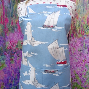 May include: A blue apron with a nautical theme, showing sailboats, lighthouses, and clouds. The apron has a pocket and a white top with a red pattern. The design is repeated across the apron's surface.