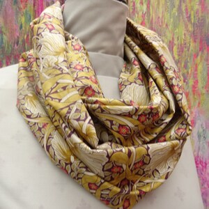May include: A silk infinity scarf with a floral pattern in shades of gold, cream, and burgundy. The design features stylized flowers and leaves. The scarf is draped around a mannequin's neck, showcasing its flowing fabric.