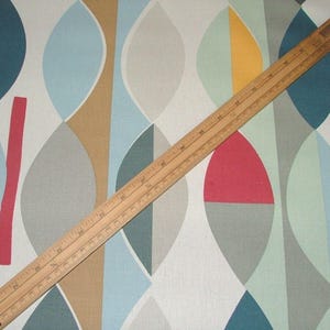 May include: A wooden ruler lies across a fabric with a colourful geometric pattern. The fabric features a repeating design of overlapping ovals in shades of blue, grey, tan, red, and yellow. The ruler is marked in inches and centimetres.