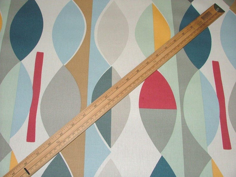 May include: A wooden ruler lies across a fabric with a colourful geometric pattern. The fabric features a repeating design of overlapping ovals in shades of blue, grey, tan, red, and yellow. The ruler is marked in inches and centimetres.