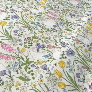 Può includere: Un tessuto color crema con un motivo ripetuto di conigli bianchi, fiori colorati e farfalle. Gli elementi floreali includono tulipani gialli, digitali rosa e iris viola. Il design ha un tema primaverile o pasquale.