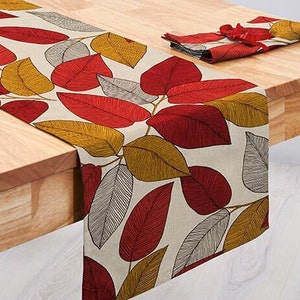 Può includere: Runner da tavolo beige con un motivo a foglie nei toni del rosso, oro e grigio. Il runner è posto su un tavolo di legno, con un set di tovaglioli abbinato. Le foglie hanno motivi di venature dettagliati.