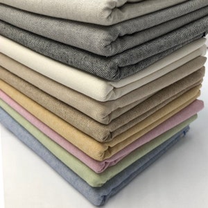 Op de afbeelding: Een stapel gevouwen stofstalen in verschillende kleuren. De stoffen variëren van lichtbeige tot donkergrijs, met extra kleuren zoals geel, roze en blauw. De stalen zijn netjes opgestapeld en tonen de verschillende texturen en tinten van het materiaal.