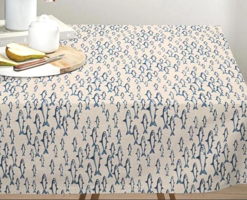 May include: A rectangular tablecloth featuring a repeating pattern of small, dark blue fish printed on a light beige fabric. The fish are oriented in various directions across the surface, creating a dense, uniform design throughout the textile.