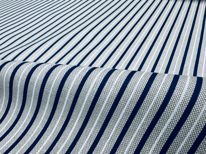 May include: A woven fabric featuring a repeating pattern of bold navy blue vertical stripes alternating with thinner white stripes against a textured light gray background. The material is folded to show the depth and weave of the textile.