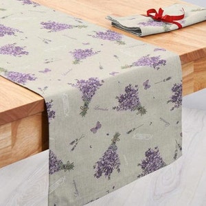 Può includere: Un runner da tavolo e un set di tovaglioli verde chiaro con un motivo floreale di lavanda. Il runner è drappeggiato su un tavolo di legno e il tovagliolo è piegato con un nastro rosso. Il design della lavanda include bouquet di fiori viola e farfalle.
