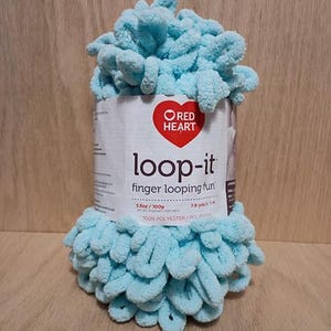 May include: A spool of light blue Red Heart Loop-It yarn. The label reads "loop-it finger looping fun". The yarn is 3.5 oz (100g) and 7.6 yds (7 m) of 100% polyester. The yarn is a soft, looped texture.
