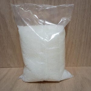May include: A clear plastic bag filled with white, granular material. The bag is sealed at the top and sits upright on a light brown wooden surface. The contents appear to be a bulk quantity of a substance, possibly for crafting or industrial use.