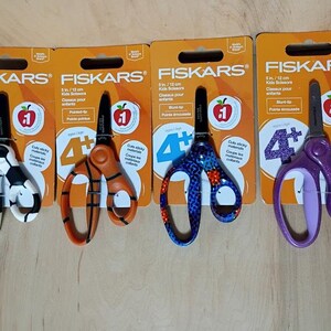 May include: Four pairs of Fiskars Kids Scissors, each on a branded orange card. The scissors have blunt tips and various handle designs: soccer ball, orange stripes, blue floral, and purple glitter. Each card has the number 4+ and an apple logo.