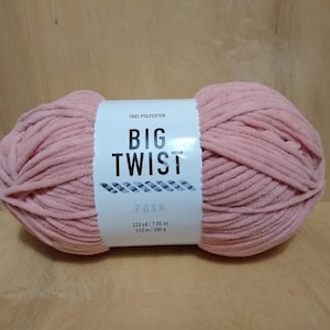 May include: A large, soft, pink yarn ball with a white label that reads "BIG TWIST POSH". The yarn is 100% polyester and measures 233 yards / 212 meters and weighs 200 grams. The yarn is on a light brown wooden surface.