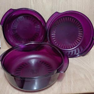 May include: Three purple plastic bowls with handles. Two of the bowls have a strainer design. The bowls are arranged on a light brown surface. The bowls are a deep, rich purple color.