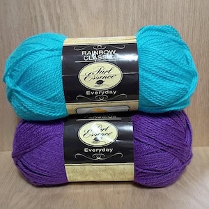 May include: Two skeins of yarn, one turquoise and one purple, are stacked on a wooden surface. Each skein has a label that reads "Rainbow Classic" and "Purl Essence Everyday."