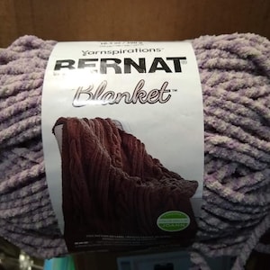 May include: A skein of Bernat Blanket yarn in a purple and white colorway. The yarn is a super bulky weight and is made of 100% polyester. The label shows a crocheted blanket in a brown colorway.