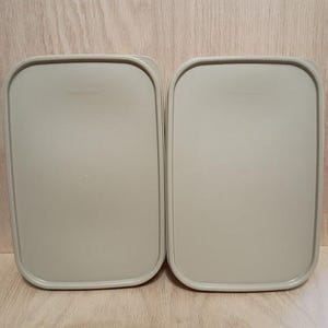 May include: Two rectangular, light beige Tupperware lids are displayed side-by-side. The lids have rounded corners and a slightly raised center. The Tupperware logo is embossed on each lid. The background is a light wood grain.