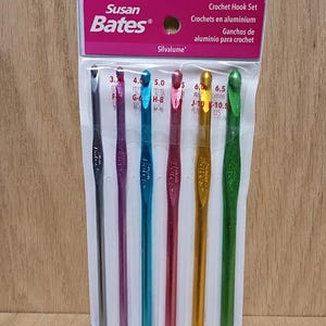 May include: A set of six crochet hooks in a clear plastic package. The hooks are in various colors, including silver, purple, teal, red, gold, and green. The package has the text "Susan Bates" and "Crochet Hook Set". The hook sizes range from 3.7 mm to 6.5 mm.