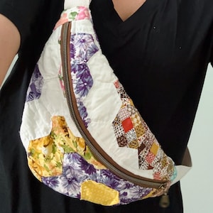Puede incluir: Un bolso bandolera acolchado con un diseño de patchwork que presenta varios patrones florales y geométricos en tonos morado, amarillo, rojo y blanco. El bolso tiene un cierre de cremallera marrón y una correa de lona ajustable en color blanco roto.