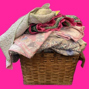 Könnte beinhalten: Ein geflochtener Korb gefüllt mit einem Stapel bunter Quilts in den Farben Rosa, Grün und Weiß. Die Quilts bestehen aus verschiedenen Stoffen und Mustern.