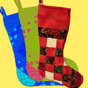 May include: Three holiday stockings featuring distinct designs. The left is blue with a geometric grid pattern. The middle is lime green with a pink diamond lattice. The right is a quilted patchwork stocking with red, cream, and floral fabric squares.