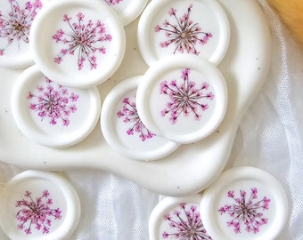 Puede incluir: Un conjunto de ocho sellos de cera blanca con flores rosas prensadas. Los sellos están dispuestos en un plato de cerámica blanca.