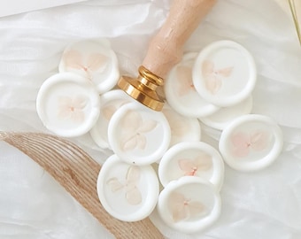 Puede incluir: Una colección de sellos de cera blanca con un delicado diseño floral en color rosa claro. Los sellos están esparcidos sobre una superficie blanca con un mango de madera y una base dorada de un sello de cera visible en el fondo.