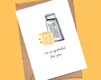 May include: A white greeting card featuring a watercolor illustration of a grey metal cheese grater and a wedge of yellow Swiss cheese with holes. The text "I'm so grateful for you" is printed in black below the illustration, resting on a brown envelope.