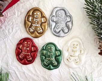 May include: A collection of five gingerbread man wax seals in gold, silver, red, green, and white. Each seal features a detailed gingerbread man design with a bow tie. The seals are arranged on a textured white surface with red ribbon and pine accents.