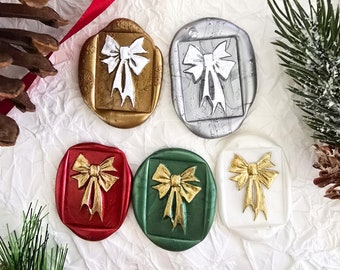 May include: Five wax seals in gold, silver, red, green, and white, each featuring a bow design. The seals are arranged on a textured white surface, with red ribbon, pine needles, and a pine cone adding a festive touch. Ideal for holiday crafts.