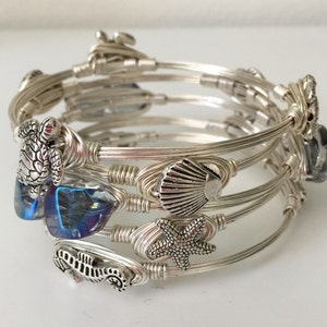 Può includere: Braccialetto avvolto in filo d'argento con ciondoli a tema marino, tra cui un cavalluccio marino, una stella marina, una conchiglia e una tartaruga. Il braccialetto include anche perline di vetro blu iridescente.