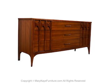 May include: A mid-century modern chest of drawers in a warm brown finish. The chest has three drawers and a decorative panel on the left side. The legs are tapered. The hardware is a dark metal. The chest is approximately 152 cm wide.