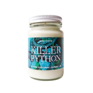 May include: A white candle jar with a green and blue label that says "Killer Python" and "Nook & Burrow". The label features a snake design and lists the scent as "Jelly Beans, Black Currant, Raspberry, Lime".