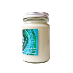May include: A white candle jar with a green and blue label that says "nook & burrow" and "since 2012" with the website "nookandburrow.com".