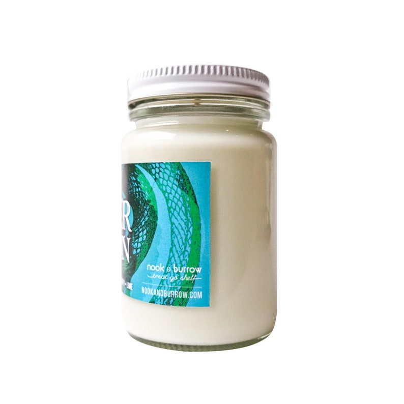 May include: A white candle jar with a green and blue label that says "nook & burrow" and "since 2012" with the website "nookandburrow.com".