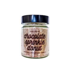 Może przedstawiać: Świeca w szklanym słoiku z czarną pokrywką. Etykieta zawiera napis "nook + burrow chocolate sprinkle donut" oraz "donut dough - chocolate glaze - sprinkles".