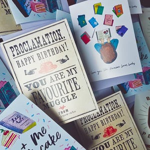 May include: A collection of greeting cards with illustrations of books, cakes, and a person reading. The cards have text that says "You are my favorite muggle" and "Books + Me + Cake".