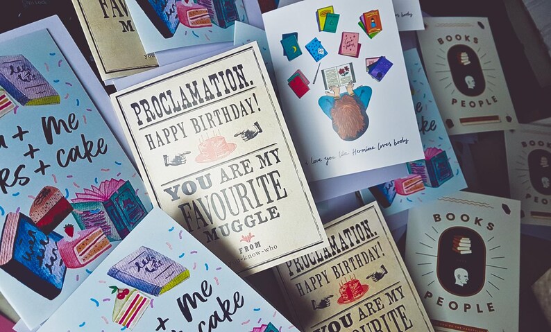 May include: A collection of greeting cards with illustrations of books, cakes, and a person reading. The cards have text that says "You are my favorite muggle" and "Books + Me + Cake".