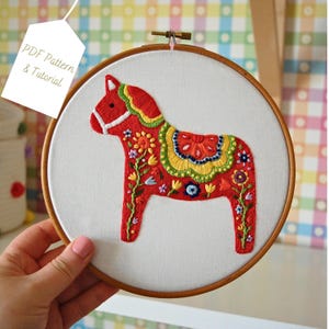May include: A red Dala horse embroidered on white fabric, set in a wooden hoop. The horse features colourful floral embroidery. A tag reads "PDF Pattern & Tutorial".