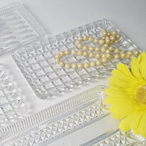 Op de afbeelding: Vier rechthoekige dienbladen van helder glas met verschillende patronen. De dienbladen zijn in een verspringend patroon op een wit oppervlak gerangschikt.