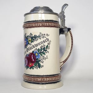 May include: A cream-coloured ceramic beer stein with a metal lid and handle. The stein features a floral design with red and blue flowers and the German text "Herzlichen Glückwunsch" printed in black script. It has decorative brown textured bands.