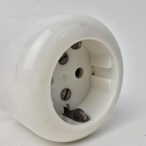 May include: A white electrical outlet with a cylindrical body. The outlet has three holes and metal screws visible. The design is simple and functional, typical of electrical components.