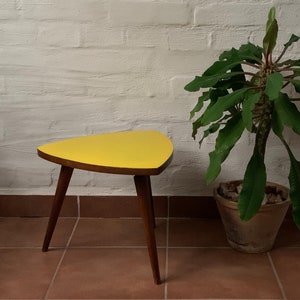 May include: A yellow triangular side table with three wooden legs. The table is sitting on a tiled floor next to a potted plant.