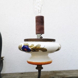 May include: A white ceramic lampshade with floral designs in shades of blue, green, and orange. The lampshade is attached to a black metal lamp base with a clear light bulb.