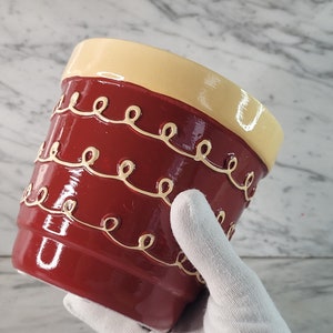 May include: A red ceramic planter with a yellow rim and a white decorative design. The planter has a glossy finish.