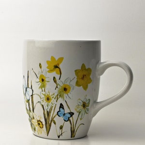 Könnte beinhalten: Weiße Keramiktasse mit einem Blumendesign mit gelben Narzissen und blauen Schmetterlingen. Die Tasse hat eine abgerundete Form und einen Henkel.