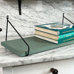 May include: A sage green floating shelf with black metal brackets, holding three books with teal covers. The shelf is mounted on a white marble surface, creating a minimalist display.
