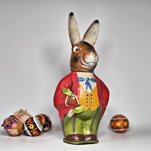Könnte beinhalten: Dekorative Osterhasenfigur mit rotem Mantel, gelber Weste und grüner Hose. Der Hase hat braunes Fell, lange Ohren und eine blaue Fliege. Mehrere dekorierte Ostereier sind um die Basis der Figur verstreut.