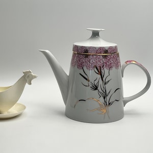 Può includere: Un set di teiera e lattiera in ceramica. La teiera è grigia con un motivo floreale bordeaux e oro. La lattiera ha la forma di un pollo ed è bianco sporco.