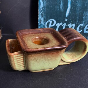 May include: A brown and green ceramic candle holder with a square top and a handle. The holder has a small, round opening in the center of the top for a candle.